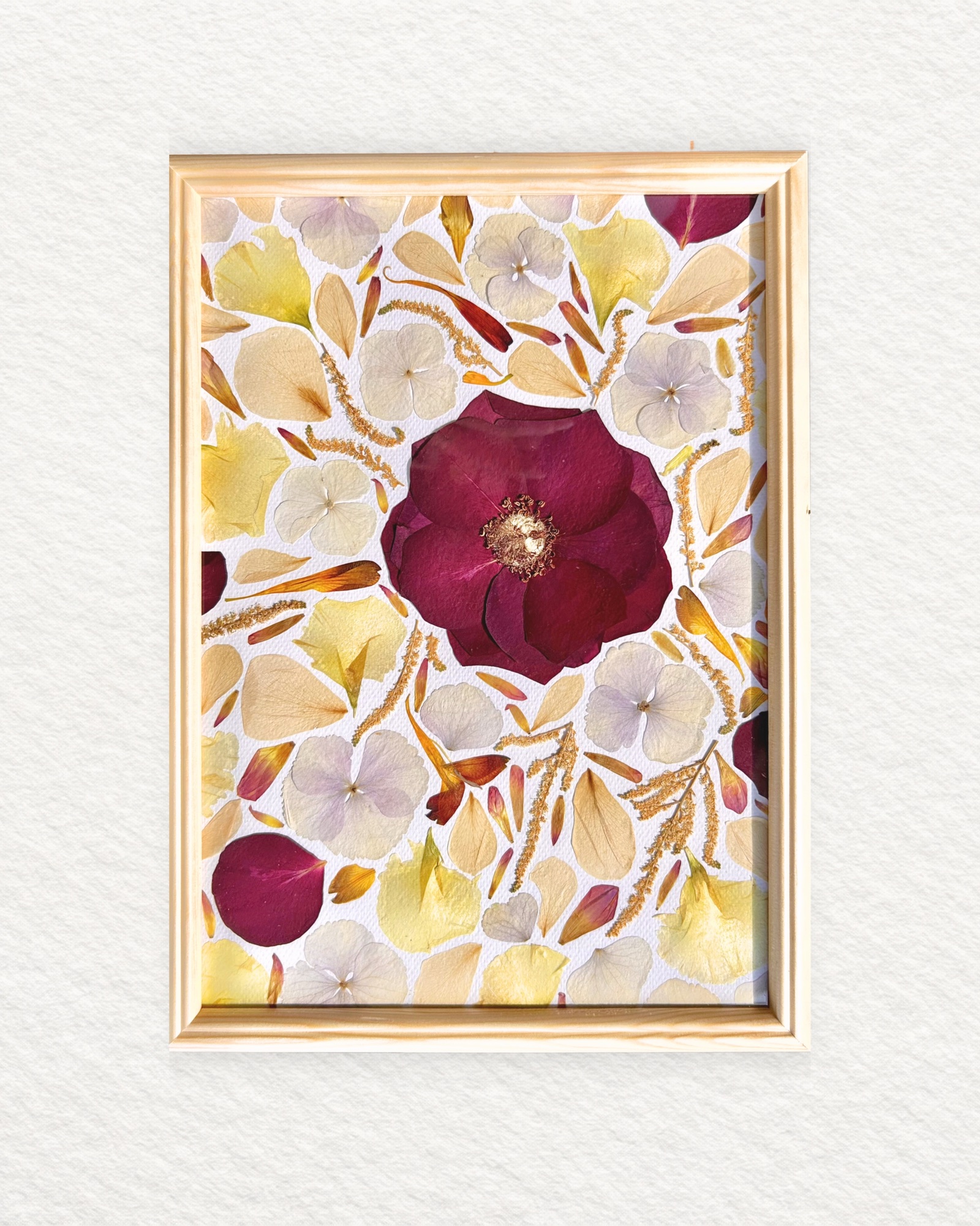 Rose rouge foncé entourée de pétales crème et jaunes, bouquet de mariée pressé, cadre en bois clair