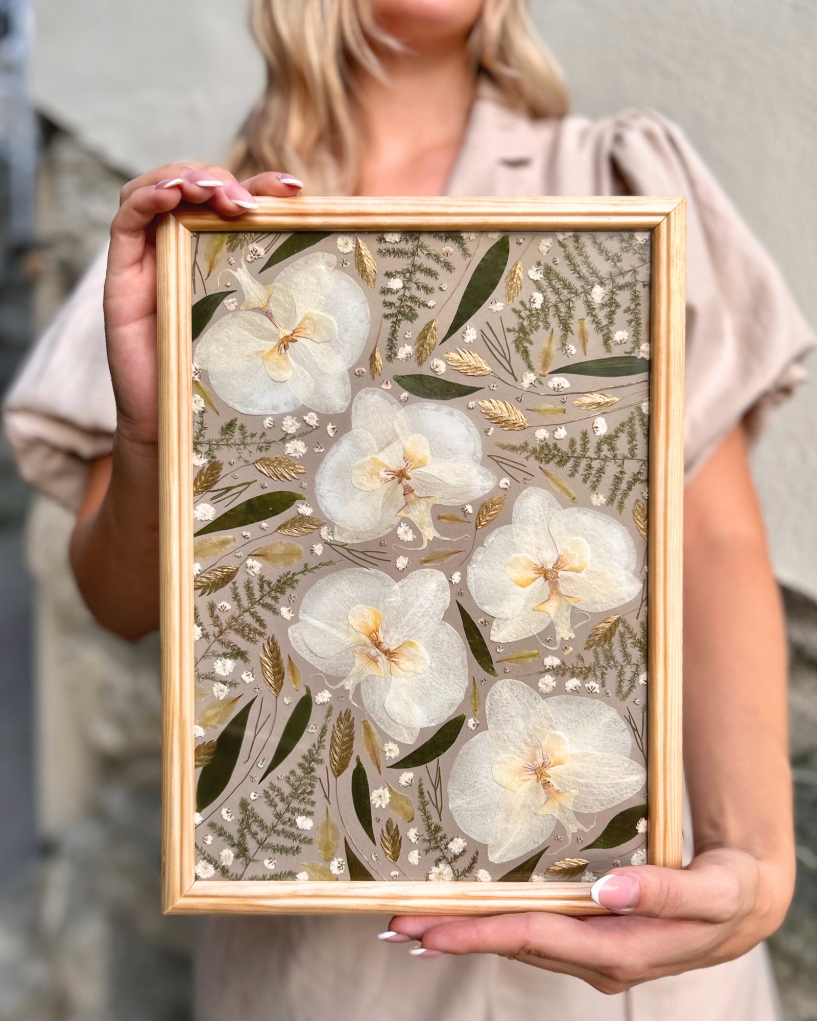 Tableau de bouquet de mariée séché : orchidées blanches, feuillage vert et blé, cadre bois clair tenu à la main